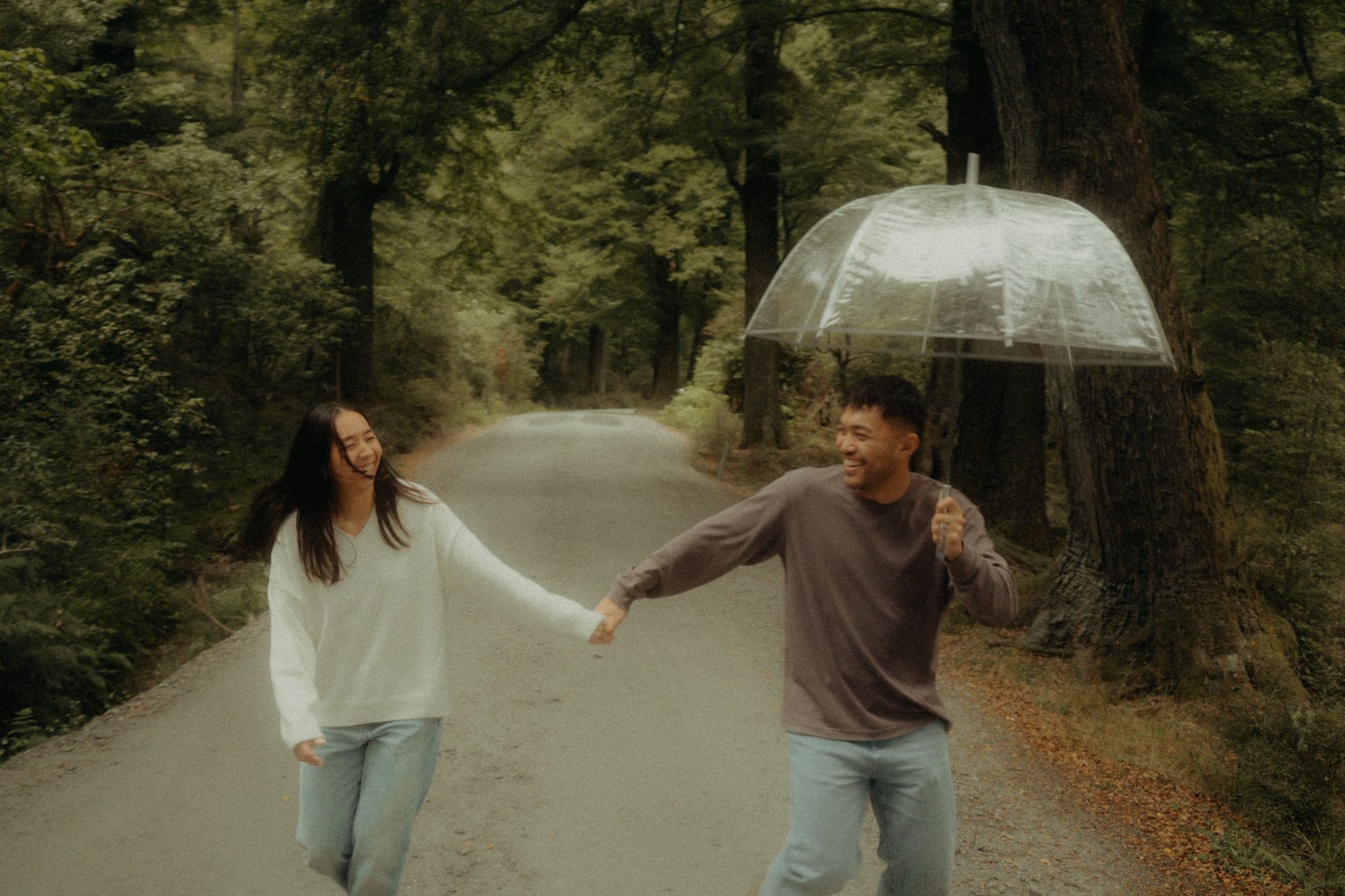 Eddison and Clarissa laughing and running hand in hand down a forest road, Eddison holding a clear umbrella